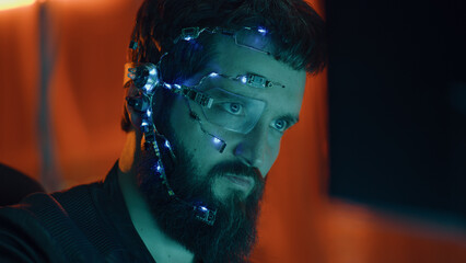 Brunette guy works on the computer with neon lights in background. Cyberpunk. Wearing a futuristic one-eyed glasses and microphone. Cyber and sci-fi backgrounds. © Framestock