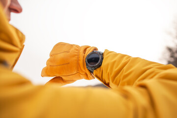 Close-up of male hands in gloves look to the watches. The man dresssed yellow warm jacket and trousers, bagpack traveling in the snow landscape