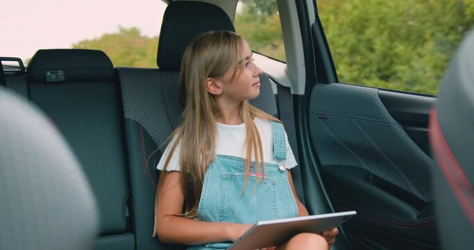 Cute positive blond teen girl sitting on car's backseat,uses her tablet pc and looking into window during trip
