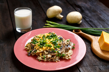 Pasta in a creamy sauce with mushrooms in a plate on the table next to a glass of cream, green onions and champignons.