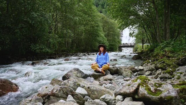 Red hair girl in a hat and blue shirtmeditates near the waterfall and the river in in the spring. Female relax outdoor