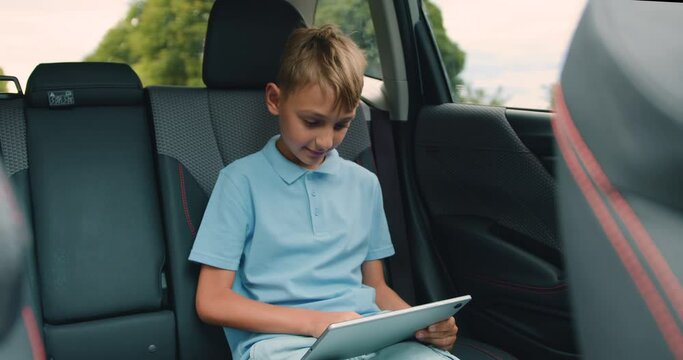 Handsome positive relaxed teen boy sitting in the car,uses his tablet pc and looking into the window from time to time during trip