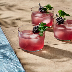 Three bramble cocktails garnished with a frozen blackberry and mint in hard light next to a pool.