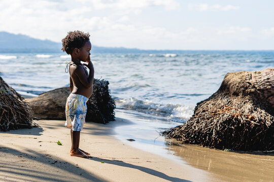 Imagen de un hermoso niño moreno afroamericano con cabello rizado y sin camisa con sus dedos en la boca en pie sobre la arena  mirando al mar de una playa en el caribe de Costa Rica.