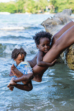 Imagen vertical  de un pequeño niño afroamericano de cabello afro  jugando abrazado a las piernas de su madre en la playa en un hermoso día de verano. 