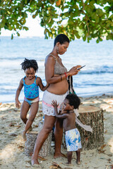 Imagen vertical de una madre africana de pie en la playa junto a sus hijos que juegan a su...