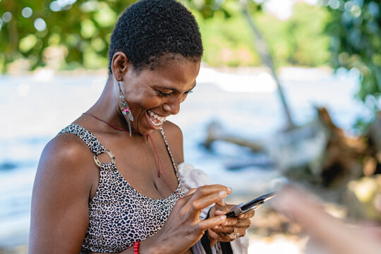 Imagen horizontal de una hermosa mujer africana de cabello corto muy sonriente sola en la playa utilizando un celular. 