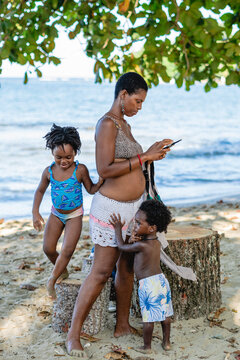 Imagen vertical de una madre africana de pie en la playa junto a sus hijos que juegan a su alrededor mientras ella utiliza el celular.