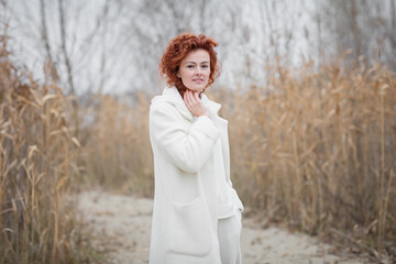 Autumn portrait. Happy redhair woman in white clothes is looking at camera and smiling while walking in the park