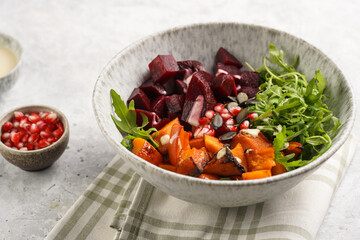 A grey deep bowl with warm winter autumn salad with arugula, red beetroot, baked pumpkin and hummus dressing cubes on a grey background