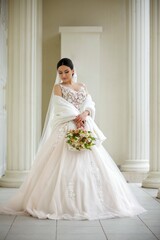 Fototapeta premium Happy beautiful young bride on the walk. Girl in a white dress with a veil. In the background are white pillars.