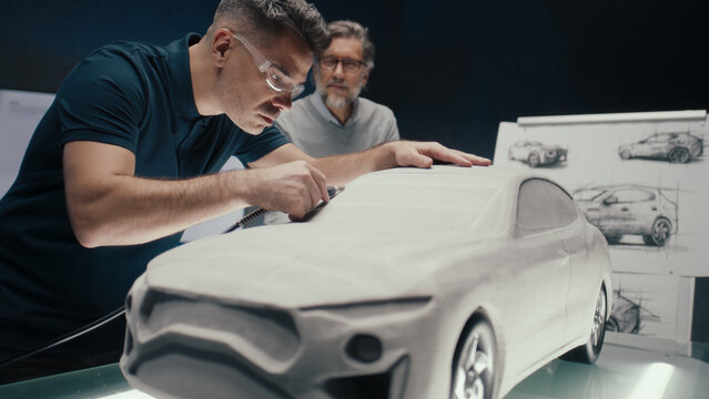 Automotive engineer works on the sculpture of a prototype car model using a rotating tool while discussing with his senior colleague. High tech car design company with innovative technologies.