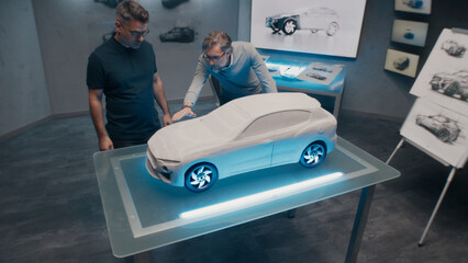 Senior automotive developers make the design corrections looking at the sculpture of the prototype car model on a glass table. High tech office with LED and car sketches.
