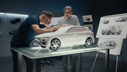 Senior automotive design engineers work on the prototype model of the car looking at the pencil sketches of the design. Using a rotary tool for final touch ups in the rake sculpture placed on a table.