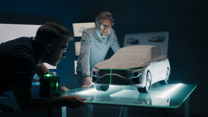Experienced automotive engineers discuss the car design infront of a sculpture prototype car model, placed on a glass table using a green laser light for alignment and perfection.