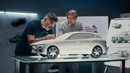 Two experienced automotive engineers discuss the car design looking at the sketches on the board. Making corrections in the prototype rake sculpture using a rotary tool with spinning tip.