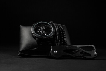 Closeup shot of a men's wristwatch with bracelets and pocket knife on black background - obrazy, fototapety, plakaty