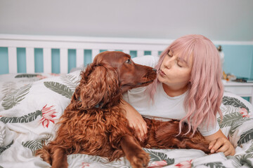 Cute pink hair woman with dog sitting in bed waking up happy concept, smiling lady awake after healthy sleep, kissing with pet