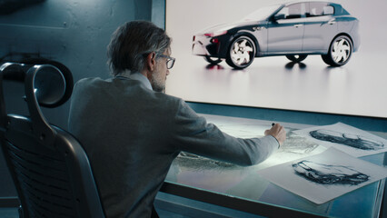 Automotive graphic designer draws sketch of the prototype model of a car with digital display graphics touch sreen table, using stylus pen in a high tech innovative laboratory.