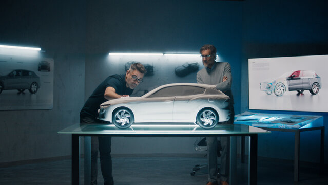 Experienced automotive engineers discuss the design infront of a rake sculpture prototype car model. Engineers use a touch screen digital tablet in a high tech laboratory. Make design corrections. - Powered by Adobe
