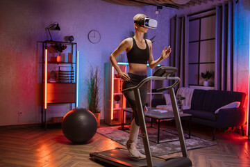 Fototapeta na wymiar Attractive young sports caucasian woman doing fitness exercise, running on treadmill using virtual reality glasses. Athletic and muscular female having actively workout indoor at night time.