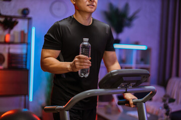 Cropped view of attractive young sports asian man with water bottle working out, running, doing cardio training on treadmill in evening time, indoor on background of modern apartment or gym.