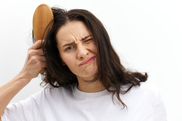 Fototapeta premium a sad, upset woman is trying to comb her long, dark, tangled hair with a wooden massage comb, standing in a white T-shirt on a white background and making a sad, funny face