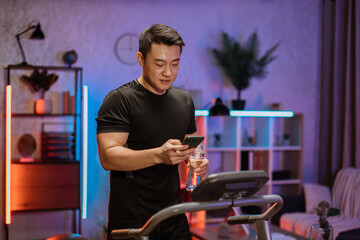 Attractive young sports asian man with water bottle using phone working out, running, doing cardio...