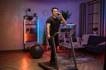 Fototapeta na wymiar Attractive young sports asian man using smartphone working out, running, doing cardio training on treadmill in evening time, indoor on background of modern apartment or gym.