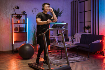 Fototapeta na wymiar Pozitive asian men in sports clothes and sneakers listening music in headphones while using smartphone, doing running exercise on electric treadmill at evening time in fitness room at home.