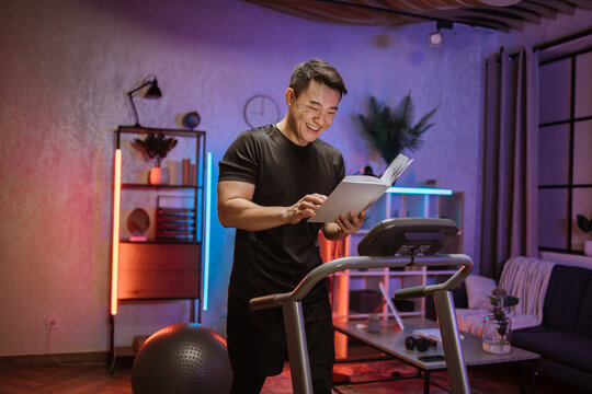 Portrait Of Handsome Active And Dynamic Young Asian Man Reading Book While Doing Sport Fitness At Home Running On Treadmill Indoor At Night. Tone Your Body. Perfect Shape. Improving Endurance.