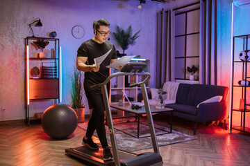 Fototapeta na wymiar Portrait of active and dynamic young handsome businessman with headset and papers doing sport fitness at home running on treadmill indoor at night. Tone your body. Perfect shape. Improving endurance