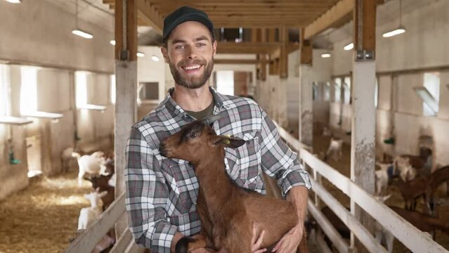 Happy young Caucasian man standing indoor at animal farm and holding baby goat in hands looking at camera and smiling. Man farmer holds brown goat in barn. Cattle in stable on background