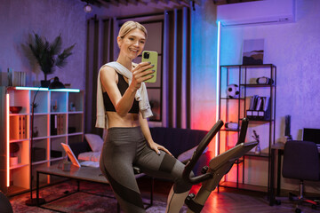 Fototapeta na wymiar Positive funny blond caucasian sports women in sportswear doing selfie on her mobile phone, cycling bike at home at night time. Cardio training, exercising legs, cardio workout indoors.