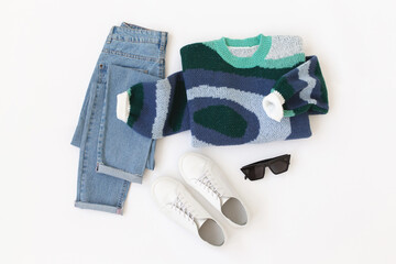Blue jeans, blue knitted sweater, white sneakers and sunglasses lying on white background. Overhead view of women's casual outfit. Trendy stylish women clothes. Flat lay, top view.