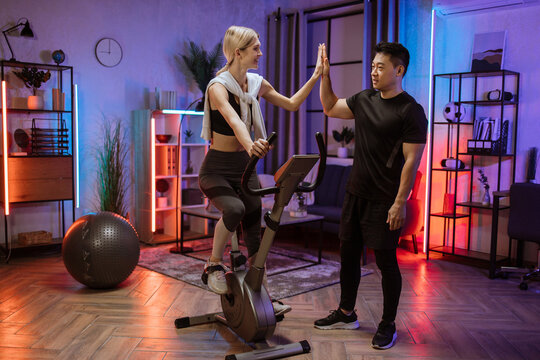 Beautiful smiling young woman doing cardio on stationary bike giving high five to her fitness trainer for good results. Attractive female working out on fitness at evening time at home .