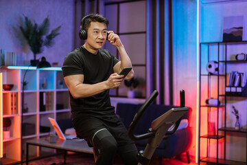 Fototapeta na wymiar Healthy fit smiling asian man training at home on exercise static bike during workout holding phone, listening music with headphones for motivation. Male healthy weekly habits app.