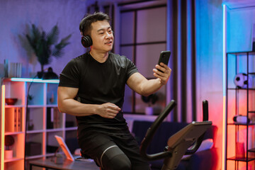 Young asian man with headphones doing exercises on stationary bicycle at home or fitness center. Young sporty male in gym listen music from smartphone. Man doing cardio exercises.