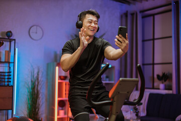 Fototapeta na wymiar Portrait of young asian male wearing sportswear having conversation on his smartphone while working out using exercise bike. Home fitness workout sporty man training on smart stationary bike indoors.