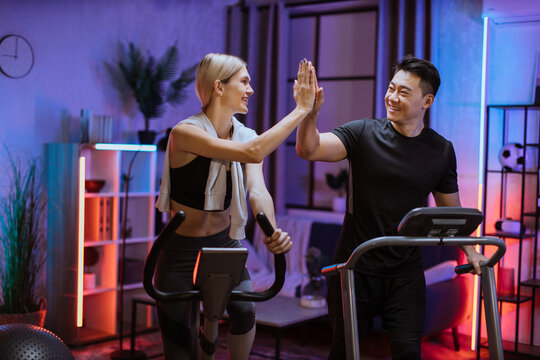 Joyful caucasian woman training on exercise bike with sporty asian man running on treadmill and giving high five. Smiling young couple doing cardio training on exercise machines at evening time