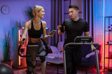 Fototapeta na wymiar Blond caucasian sports women in sportswear cycling bike and fit asian man running on treadmill and showing each other photos from social networks.