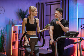 Young couple doing cardio on stationary bike and treadmill using smart phones to control physical parameters. Attractive female and male working out on fitness at evening time at home.