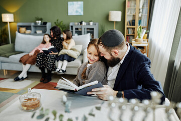 Portrait of smiling jewish father reading book to daughter while enjoying family time at home, copy...