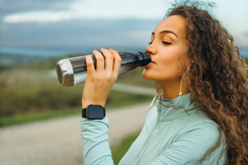 A girl is drinking water from her transparent bottle, a smart watch on her wrist, and headphones in her ears.