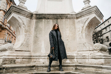 Front view of confident woman in stylish outfit posing near part of old building. Fashionable girl with brown long hair, dressed in quilted down jacket and holding bag, standing in ancient town - obrazy, fototapety, plakaty