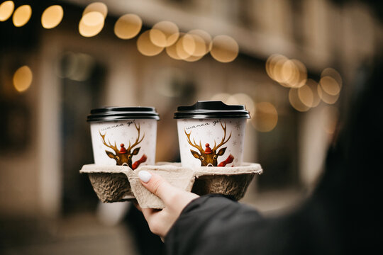 Paper cups of hot beverage in special vessel on background of Christmas lights. Incognito person, holding a cups of coffee with pictures of deer against of bokeh from garlands in decorated city street