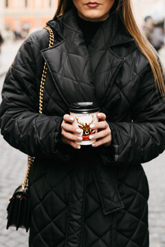 Crop of woman in fashionable apparel holding paper cup of coffee. Faceless girl with white manicure, dressed in quilted warm black jacket, heating hands to a paper cup of tea during walk outdoor