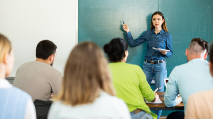 Young woman teacher is giving a lecture to students, standing near the blackboard in the university...
