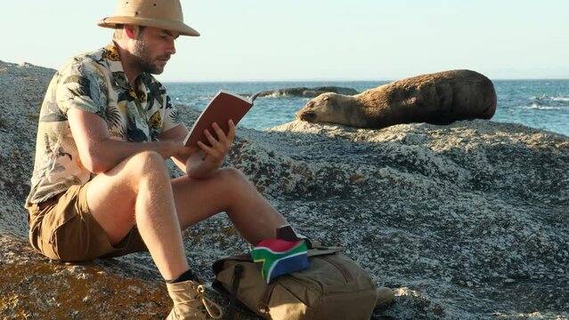 a male traveler in a safari hat stands near a young seal on the ocean. a male photographer takes a photo on the sea beach of a sleeping seal in the sun . man reading a book on the beach south africa