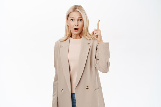 Image of mature blond woman looking amazed, saying wow, pointing finger up, eureka gesture, standing over white background